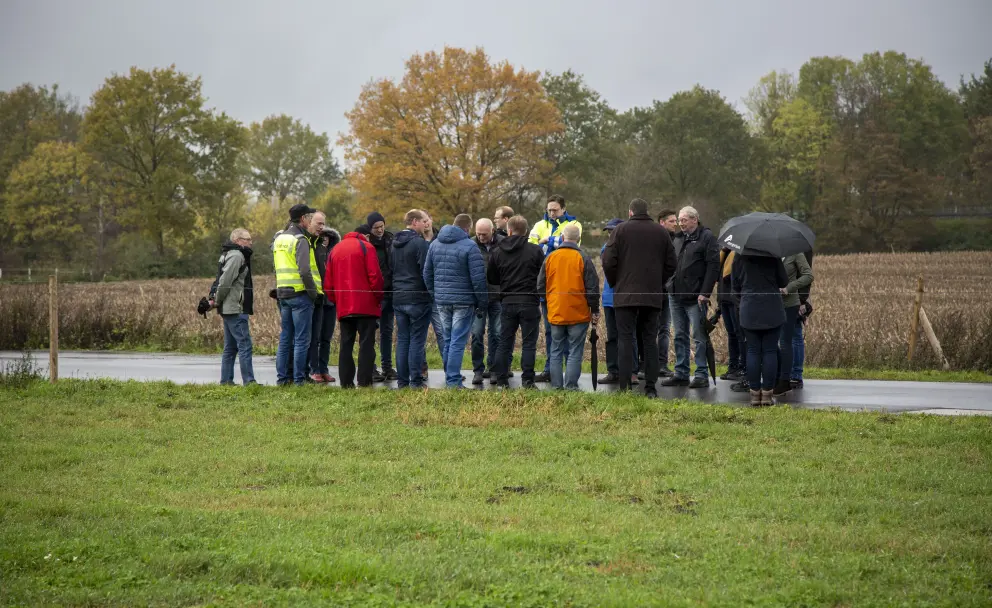 Eine Außenaufnahme, die eine Gruppe von etwa 15 Männern auf einem schmalen, asphaltierten Weg zeigt, der an eine grüne Wiese und ein dunkles, gepflügtes Feld angrenzt. Sie stehen in einer lockeren Formation beieinander und scheinen einem unsichtbaren Sprecher im Zentrum der Gruppe zuzuhören. Die Personen tragen legere, wettergerechte Kleidung in dunklen Farben wie Schwarz, Blau und Grau, ergänzt durch einige leuchtende Akzente wie eine rote Jacke, eine gelbe Warnweste und eine orangefarbene Weste. Eine Person am rechten Rand schützt sich mit einem schwarzen Regenschirm. Der Himmel ist bedeckt und grau. Im Hintergrund erstreckt sich eine offene, grüne Landschaft mit Bäumen in der Ferne.
Kontext der Szene