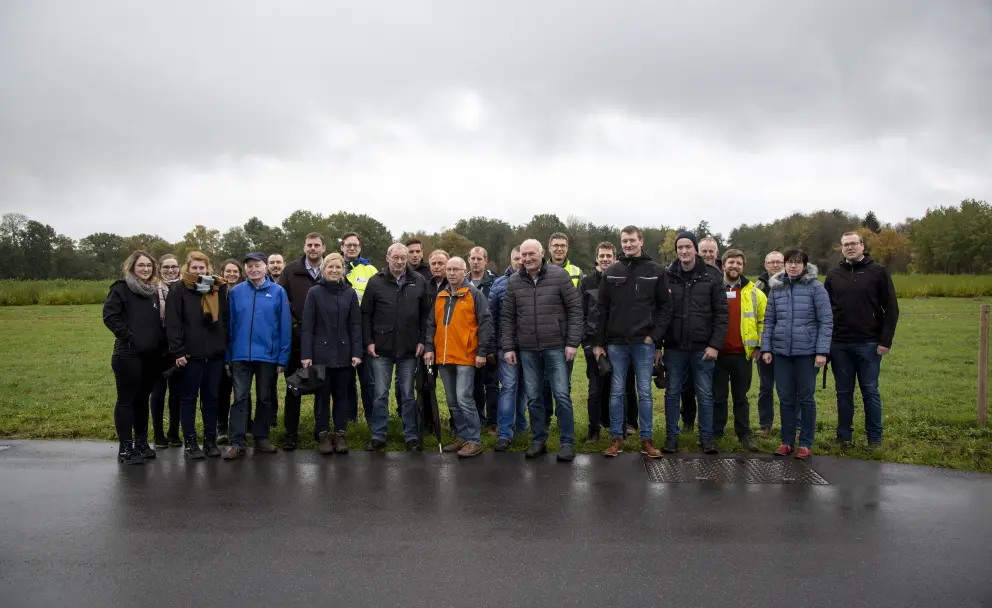 Eine frontale Außenaufnahme einer großen Gruppe von etwa 20 Erwachsenen, die für ein Foto aufgereiht sind. Sie stehen auf einer nassen, asphaltierten Fläche, die an eine grüne Wiese grenzt, unter einem bewölkten, grauen Himmel. Die Personen tragen legere Freizeit- und Arbeitskleidung, die dem kühlen Wetter entspricht, darunter Jacken, Mäntel und Jeans in verschiedenen dunklen Farben sowie einige helle Akzente wie eine orangefarbene und eine gelbe Jacke. Der Hintergrund zeigt eine offene, grüne Feldlandschaft mit dichter Baumbewachs in der Ferne. Alle Personen blicken in die Kamera.