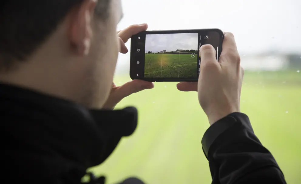 Eine Nahaufnahme aus der Rückenperspektive zeigt einen Mann in einem schwarzen Oberteil, der mit beiden Händen ein schwarzes Smartphone im Querformat hält, um eine Aufnahme zu machen. Der Bildschirm des Telefons zeigt die Kamera-Benutzeroberfläche und die Live-Vorschau einer Landschaft mit einem grünen Feld und einer gepflasterten Fläche im Vordergrund. Die Perspektive des Bildschirms ist fast identisch mit der des Hauptfotos. Die Ränder des Telefons sind leicht abgerundet und das Display ist hell beleuchtet. Der Rest des Bildes, insbesondere die reale grüne Umgebung um das Telefon herum, ist unscharf gehalten.
