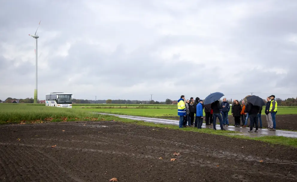 Eine Außenaufnahme bei bedecktem Himmel zeigt eine Gruppe von etwa einem Dutzend Personen auf einem unbefestigten, matschigen Feldweg. Links im Bild steht, von hinten gesehen, eine Person mit einer auffälligen gelben Warnweste. Die Gruppe steht in einem Kreis beisammen, einige Personen schützen sich mit dunklen Regenschirmen vor leichtem Regen. Im Hintergrund links ist ein hoher, weißer Windkraftgenerator mit einem orangefarbenen Streifen auf dem Turm und einem grünen Farbakzent an der Gondel zu sehen. Rechts neben dem Windrad steht ein weißer Reisebus auf der angrenzenden Wiese. Der Boden im Vordergrund ist ein gepflügtes, dunkles Feld, und im Hintergrund erstreckt sich eine weite, grüne Feldlandschaft mit Bäumen in der Ferne.