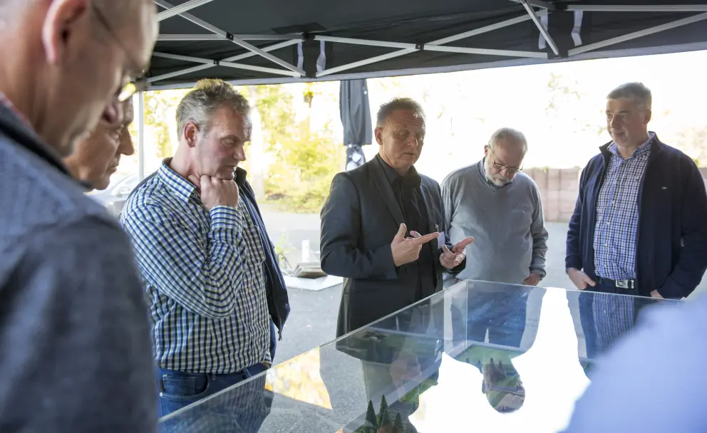 Eine Nahaufnahme aus der Vogelperspektive zeigt eine Gruppe von fünf Männern, die unter einem schwarzen Pavillonzelt stehen und ein gläsernes Tischmodell betrachten. Der Mann in der Mitte trägt einen schwarzen Anzug und gestikuliert mit beiden Zeigefingern in Richtung des Modells. Um ihn herum stehen andere Männer in legerer Freizeitkleidung, darunter karierte Hemden, Pullover und Jeans. Sie blicken alle nach unten auf das Objekt. Im Hintergrund sind unscharf grüne Bäume und ein beigefarbener Zaun zu sehen.
