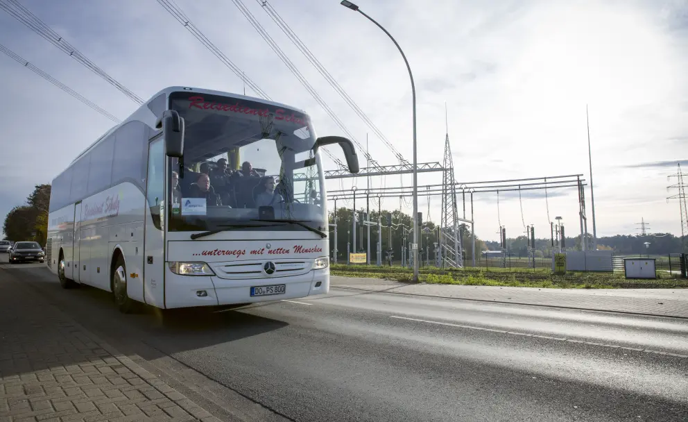 Eine frontale Außenaufnahme eines großen, weißen Reisebusses der Marke Mercedes-Benz Tourismo auf einer befestigten Straße bei hellem Tageslicht. Auf der Front und Seite des Busses ist der rote Schriftzug "...unterwegs mit netten Menschen" zu lesen. Das Kennzeichen DO PS 600 ist vorne sichtbar. Im Hintergrund rechts befindet sich eine komplexe Elektro-Umspannstation mit mehreren Stahlmasten und Isolatoren. Links hinter dem Bus sind weitere Fahrzeuge und Bäume zu sehen.
