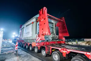 Ein roter Wagenborg Nedlift Schwerlast-LKW-Zug fährt nachts auf einer nassen, asphaltierten Straße. Der LKW ist ein MAN TGX, der einen großen, hellgrauen Transformator auf einem mehrachsigen, modularen Tieflader transportiert. 
Die Szene wird durch helle Straßenlaternen und die eingeschalteten Lichter des LKW beleuchtet. Im Hintergrund ist ein zweistöckiges, giebelständiges Gebäude mit beleuchteten Fenstern und einem Amprion-Logo im Fenster zu sehen. 
