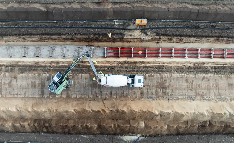 Eine Luftaufnahme zeigt eine Baustelle in einem offenen Grabenbereich.
Oberer Bereich: Dunkles Erdreich bildet den oberen Rand. Eine vorbereitete, ebene Fläche mit feinem, grauem Material erstreckt sich horizontal. Schwarze, schmale Rohre verlaufen quer im Erdreich, und ein kleiner, orangefarbener Kasten ist sichtbar.
Mittlerer Bereich: Eine graugrüne Baumaschine mit einem langen, knickbaren Arm steht auf der linken Seite der vorbereiteten Fläche. Rechts daneben steht ein weißer Betonmischer-LKW. Eine schmale Rutsche verbindet den Mischer mit einem vorderen Bereich. In diesem vorderen Bereich werden rote Rohre mit hellem Material (vermutlich Beton) ummantelt. Zwei Personen in orangefarbener Warnkleidung sind in der Nähe der Maschinen und der Rohre zu sehen.
Unterer Bereich: Ein breiterer, tieferer Graben mit losem, sandigem braunem Aushub nimmt den unteren Teil des Bildes ein. Im untersten Rand sind weitere schwarze Rohre im Boden erkennbar.