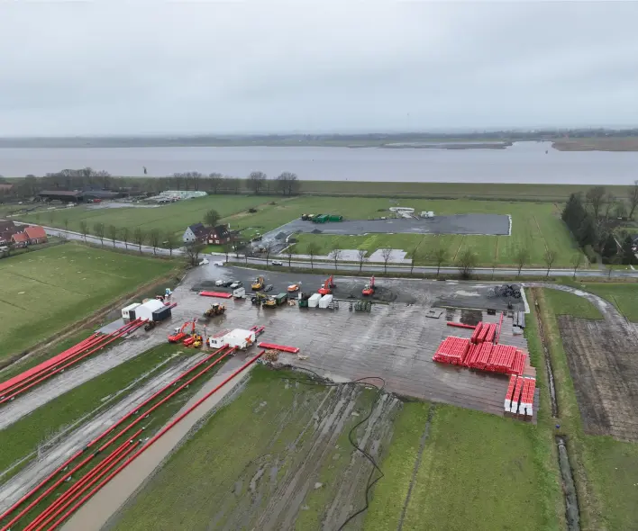 Eine Luftaufnahme zeigt ein ländliches Gebiet neben einem größeren Gewässer mit einer Fläche, auf der industrielle Materialien gelagert werden.
Vordergrund: Die untere Hälfte des Bildes besteht aus grünen Wiesen, teils matschig, und einer grauen, befestigten Fläche. Auf dieser Fläche liegen mehrere große, gestapelte Haufen von roten Rohren. Links davon verlaufen weitere lange, rote Rohre in parallelen Reihen über das nasse Gras. Es sind auch mehrere gelbe und orangefarbene Maschinen (Bagger, Radlader) sowie einige weiße und schwarze Container oder Zelte sichtbar.
Mitte: Die befestigte Fläche geht in eine asphaltierte Straße über, die durch eine Reihe von Bäumen gesäumt ist. Hinter der Straße liegen weitere grüne Felder und ein kleineres Areal mit gelagertem grünem und weißem Material.
Hintergrund: Ein großer Erdwall oder Deich, bewachsen mit Gras, erstreckt sich horizontal im oberen Bereich des Bildes. Dahinter liegt eine breite Fläche Wasser (Flussmündung oder Meer), die in der Ferne auf einen hellgrauen, bewölkten Himmel trifft. Am linken und rechten Rand sind einige rote Backsteinhäuser mit dunklen Dächern erkennbar.