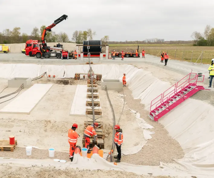 Vordergrund: Die untere Bildhälfte zeigt eine große, ausgehobene Grube mit sandigem Erdreich, die teilweise mit weißer Plane ausgelegt ist. Mehrere Arbeiter in orangefarbener Warnschutzkleidung und Helmen sind im vorderen Grubenbereich bei einer vertikalen Metallkonstruktion tätig. Auf der linken Seite liegen rechteckige Betonplatten und schwarze, schlangenartige Kabel oder Schläuche. Rote Rohrsegmente liegen ebenfalls am unteren Rand.
Mitte: Am oberen Rand der Grube steht ein roter Lastwagen mit einem montierten Kranarm. Daneben befinden sich eine schwarze Containerstruktur, eine Kabelrolle und weitere Baugeräte. Auf der rechten Seite führt eine rote Metalltreppe in die Grube hinab.
Hintergrund: Im Hintergrund sind weitere Arbeiter, weiße und gelbe Lieferwagen sowie eine Gruppe von Bäumen und offene Felder unter einem bedeckten Himmel sichtbar.
