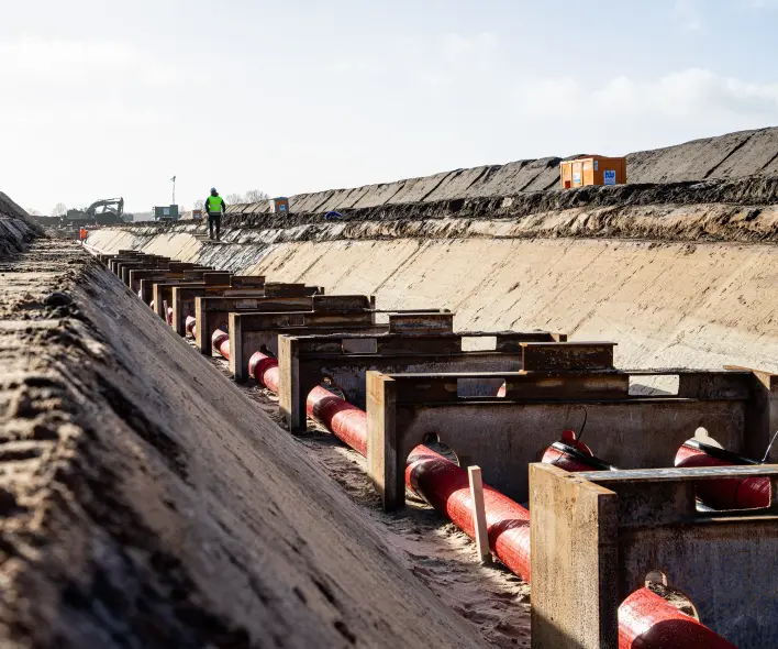 Eine Szene in einem offenen Graben zeigt die Verlegung von roten Rohren in speziellen Halterungen.
Vordergrund: Die unteren Ränder des Bildes bestehen aus rohem, losem Erdreich. Eine Reihe von rechteckigen, metall- und betonummantelten Halterungen verläuft in einer diagonalen Linie durch das Bild. In diesen Halterungen sind dicke, rote Rohre fixiert.
Mitte: Die Wände des Grabens sind hoch und bestehen aus abgeflachtem, hellem Bodenmaterial. Im oberen Bereich der linken Wand sind gestapelte Schichten dunkleren Erdreichs zu sehen. Auf dem oberen Rand des Grabens, in der Ferne, steht eine Person in einer gelben Weste und Helm. Dahinter sind weitere Baumaschinen (Bagger) und kleine Container sichtbar.
Hintergrund: Der Horizont zeigt einen hellen, teilweise überbelichteten Himmel.
