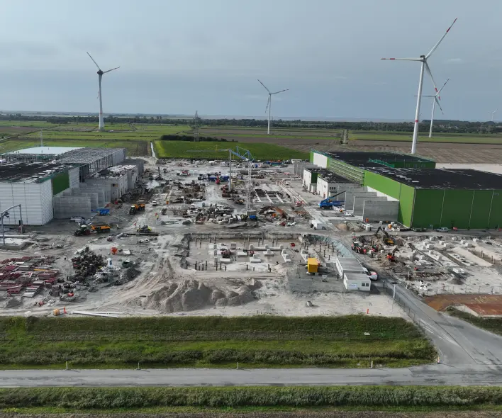 Vordergrund und Umgebung
Im direkten Vordergrund verläuft eine asphaltierte Straße mit einer durchgezogenen weißen Linie.
Eine grasbewachsene Böschung trennt die Straße von der Baustelle.
Ein grauer Zaun mit einem orangefarbenen Schild grenzt das Gelände ab.
Rechts unten führt eine weitere asphaltierte Zufahrt auf das Gelände.
Baustelle und Gebäude
Das zentrale Areal ist eine ausgedehnte, flache Baustelle mit losem, hellem Erdreich und Sand.
Mehrere Gebäudestrukturen befinden sich im Bau, mit weißen Fassaden und teilweise schwarzen Dächern (links und rechts).
Die Gebäude auf der rechten Seite haben markante grüne Fassadenelemente.
Zahlreiche Baumaschinen (Bagger, Radlader) in Gelb, Orange und Blau sind auf dem Gelände verteilt.
Im linken Mittelgrund lagern stapelweise rote und graue Baumaterialien.
Fundamente aus Beton und sichtbare Stahlbewehrungen sind im zentralen Bereich der Baustelle erkennbar.
Mehrere weiße Container oder Bauwagen sind auf der rechten Seite der Baustelle positioniert.
Hintergrund
Die Baustelle ist umgeben von grünen Wiesen und Ackerland.
Mehrere hohe Windkraftanlagen mit weißen Türmen und roten Markierungen an den Rotorblättern sind in der Ferne sichtbar.
Im linken Hintergrund sind weitere Felder und eine flache Landschaft unter einem bedeckten Himmel mit grauen und weißen Wolken erkennbar.
