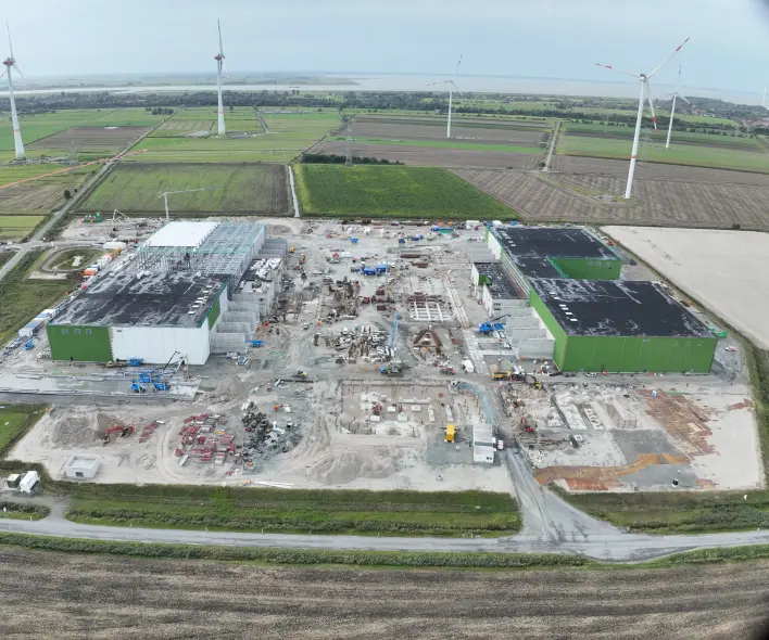 Eine Luftaufnahme zeigt eine große Baustelle in einer ländlichen Umgebung mit mehreren Windkraftanlagen im Hintergrund.
Vordergrund: Eine schmale, asphaltierte Straße verläuft horizontal. Dahinter folgt eine Böschung mit trockenem Gras und Erde.
Mitte: Eine große Baustelle nimmt den zentralen Bereich ein. Links steht ein großes Gebäude im Rohbau mit weißer Fassade und freiliegender schwarzer Dachstruktur. Rechts ein ähnliches Gebäude, ebenfalls im Rohbau, mit weißen Wänden und teilweise installierten grünen Wandpaneelen an den oberen Abschnitten. Zahlreiche Baumaschinen in Gelb, Blau und Orange, Betonfundamente, gelagertes Material (rote Rohre, graue Blöcke) und lose Erde sind auf dem Gelände verteilt.
Hintergrund: Die Baustelle grenzt an landwirtschaftlich genutzte Felder (grüne und braune Ackerflächen), die durch Wege unterteilt sind. Mehrere hohe Windkraftanlagen sind über die Landschaft verteilt. Am fernen Horizont ist eine Wasserfläche unter einem hellgrauen, bewölkten Himmel erkennbar.
