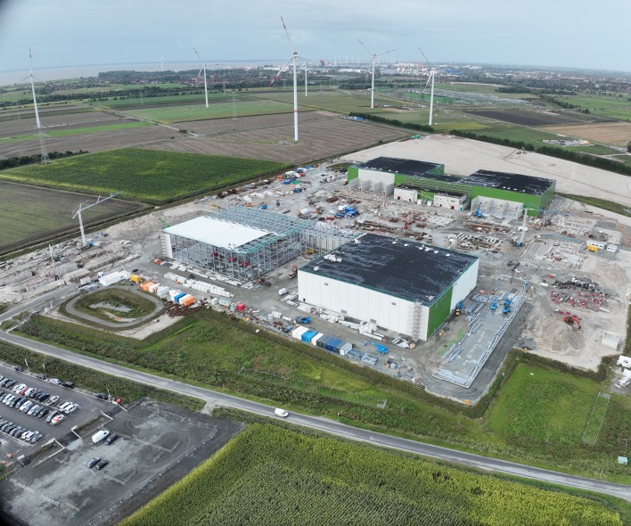 Eine Luftaufnahme zeigt eine große Industriebaustelle in einer ländlichen Umgebung.
Vordergrund und Umgebung
Im direkten Vordergrund befindet sich ein Parkplatz mit mehreren geparkten Autos.
Eine asphaltierte Straße mit weißer Markierung verläuft horizontal über das Bild und trennt den Parkplatz von der Baustelle.
Grünflächen (Wiesen und Felder) umgeben die gesamte Baustelle.
Baustelle und Gebäude
Die zentrale Baustelle ist ein großes, flaches Areal mit viel losem, hellem Erdreich und Sand.
Mehrere große Gebäude sind im Bau:
Links ein Gebäude im Rohbau mit weißer Dachstruktur und freiliegendem Stahlgerüst.
Rechts ein großes, geschlossenes Gebäude mit weiß-grüner Fassade und schwarzem Flachdach.
Im oberen Bereich weitere verbundene Gebäude mit grünen Fassaden und schwarzen Dächern.
Zahlreiche Baumaschinen in Gelb, Orange und Blau, gestapelte Materialien (Container, Paletten, Rohre) und Betonfundamente sind auf dem Gelände verteilt.
Mehrere Krane, darunter ein großer weißer Gittermastkran links, sind sichtbar.
Hintergrund
Die Baustelle grenzt an landwirtschaftlich genutzte Felder (grüne und braune Ackerflächen).
Zahlreiche hohe Windkraftanlagen mit weißen Türmen und roten Markierungen an den Rotorblättern sind in der Ferne über die Landschaft verteilt.
Am fernen Horizont ist eine Wasserfläche unter einem bedeckten Himmel mit grauen und weißen Wolken erkennbar.