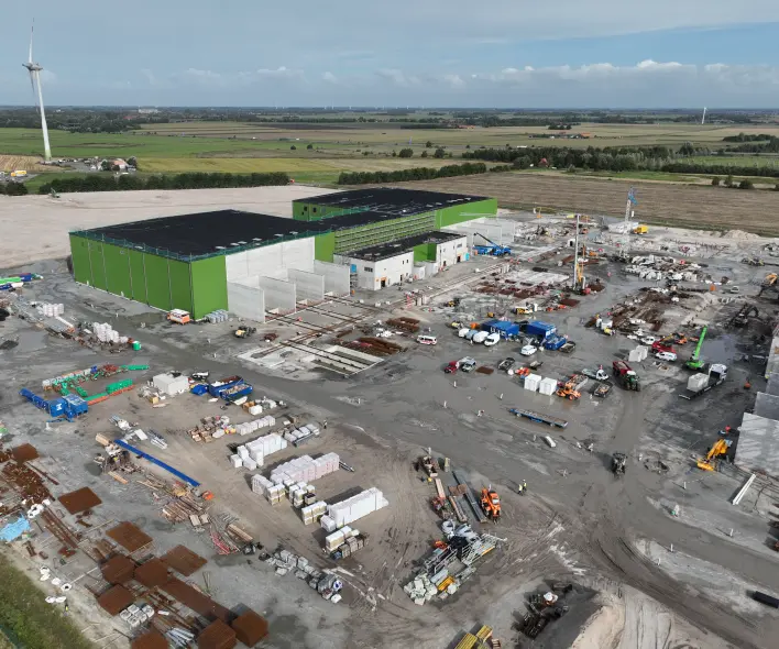 Eine Luftaufnahme zeigt eine große Industriebaustelle in einer ländlichen Umgebung.
Vordergrund: Die untere Hälfte des Bildes besteht aus losem, braunem Erdreich, Baustraßen und gelagerten Baumaterialien. Dazu gehören Bündel von Bewehrungsstahl, Betonfertigteile, rote Rohre, Paletten mit weißen Materialien, sowie verschiedene Baumaschinen (Bagger, Radlader) in Gelb, Orange und Blau. Mehrere weiße und graue Container sind ebenfalls sichtbar.
Mitte: Im Zentrum der Szene stehen mehrere große, im Bau befindliche Industriegebäude. Das Hauptgebäude links hat eine markante grüne Fassade und ein schwarzes Flachdach. Es ist mit kleineren Gebäudeeinheiten in Weiß und Grau verbunden, die ebenfalls teilweise grüne Fassadenbereiche aufweisen. Betonfundamente und freiliegende Stahlstrukturen sind im zentralen Baubereich erkennbar.
Hintergrund: Die Baustelle ist umgeben von grünen Wiesen und braunen Ackerflächen, die durch schmale Wege und eine asphaltierte Straße im oberen linken Bereich unterteilt sind. Mehrere hohe, weiße Windkraftanlagen mit roten Markierungen an den Rotorblättern sind in der Ferne sichtbar. Der Himmel ist bedeckt mit grauen und weißen Wolken.
