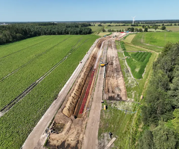 Eine Luftaufnahme zeigt eine große Baustelle, die sich diagonal durch eine ländliche Landschaft mit grünen Feldern und Wäldern erstreckt.
Vordergrund und Mitte: Ein breiter, geräumter Baustreifen verläuft von links unten nach rechts oben. Der Streifen besteht aus hellem, unbewachsenem Erdreich und Baustraßen. Entlang des linken Randes des Streifens liegt ein tiefer Graben, in dem mehrere parallele Reihen von roten Rohren liegen, die teilweise mit Erde bedeckt sind. Ein gelber Bagger ist im zentralen Bereich des Streifens positioniert. Auf der rechten Seite des Streifens befindet sich ein kleinerer Grabenbereich und Haufen von dunklerem, feuchtem Erdreich.
Hintergrund: Der Baustreifen führt in eine Landschaft mit weiteren Ackerflächen, einem großen Waldstück auf der rechten Seite und einem kleineren Wald auf der linken Seite. In der Ferne, leicht rechts von der Mitte, steht eine einzelne Windkraftanlage. Der Himmel ist hellblau mit einigen weißen Wolken.