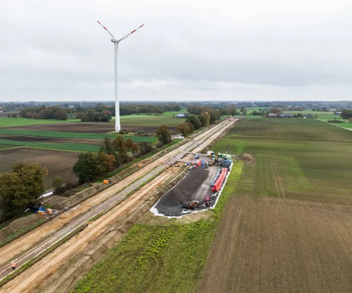 Eine Luftaufnahme zeigt eine große Baustelle in einer ländlichen Umgebung mit mehreren Windkraftanlagen im Hintergrund.
Vordergrund und Mitte
Ein breiter, geräumter Baustreifen erstreckt sich diagonal von links unten nach rechts oben durch die Landschaft.
Der Streifen besteht aus hellem, unbewachsenem Erdreich und ist durch schmale, sandige Baustraßen strukturiert.
Im rechten Mittelgrund befindet sich ein Bereich mit einem Haufen aus dunklem Material (Erde/Schotter) auf weißer Folie.
Mehrere Baumaschinen in Gelb, Blau und Orange, Baustellenfahrzeuge (grau, rot, blau) und gelagerte Baumaterialien (rote Rohre, Paletten, Container) sind in diesem Bereich angeordnet.
Links vom Hauptstreifen befindet sich ein schmaler Graben mit einigen blauen Planen.
Ein kleiner Arbeiter in orangefarbener Warnkleidung ist im unteren linken Bereich des Streifens sichtbar.