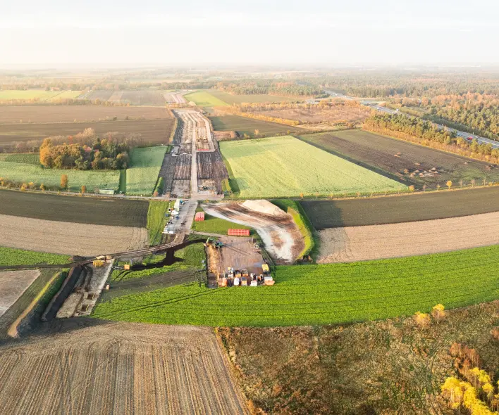 Eine Luftaufnahme zeigt eine große Baustelle, die sich diagonal durch eine ländliche, landwirtschaftliche Landschaft erstreckt.
Vordergrund und Mitte: Der untere Teil des Bildes besteht aus braunen, gepflügten Feldern und grünen Ackerflächen. Eine breite, geräumte Baustraße mit hellem Untergrund verläuft von links unten nach rechts oben. Entlang dieser Trasse liegen mehrere lange Reihen paralleler roter Rohre. Einige kürzere, leuchtend rote Rohre und gelbe/weiße Baumaschinen sind im unteren rechten Bereich sichtbar.
Hintergrund: Dahinter liegen weitere Felder in verschiedenen Braun- und Grüntönen sowie ein großer Wald mit gelbem, orangefarbenem und grünem Laub. Am Horizont ist eine flache, offene Landschaft unter einem hellen, leicht bewölkten Himmel zu sehen.