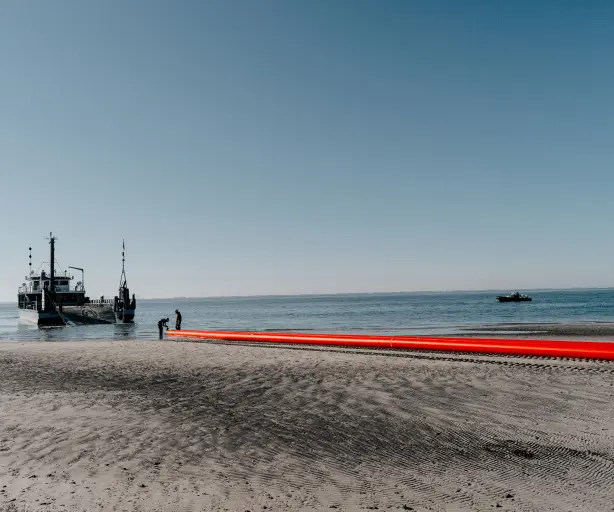 
Eine Szene am Strand, aufgenommen auf Augenhöhe, zeigt einen schwarzen Lastkahn und eine orangefarbene Rohrleitung im flachen Wasser nahe der Küste.
Vordergrund: Der untere Teil des Bildes besteht aus nassem, strukturiertem Sand.
Mitte: Im flachen Wasser auf der rechten Seite arbeiten zwei Personen an einem langen, leuchtend orangefarbenen, schwimmenden Objekt, das wie eine Pipeline oder ein Schlauch aussieht und ins Wasser ragt. Auf der linken Seite liegt ein grauer und schwarzer Lastkahn mit einer offenen Rampe am Heck vor Anker.
Hintergrund: Das ruhige, blaue Meer erstreckt sich bis zum Horizont, wo es auf einen klaren, hellblauen Himmel trifft. Links im Hintergrund ist ein kleiner Segelbootmast sichtbar.