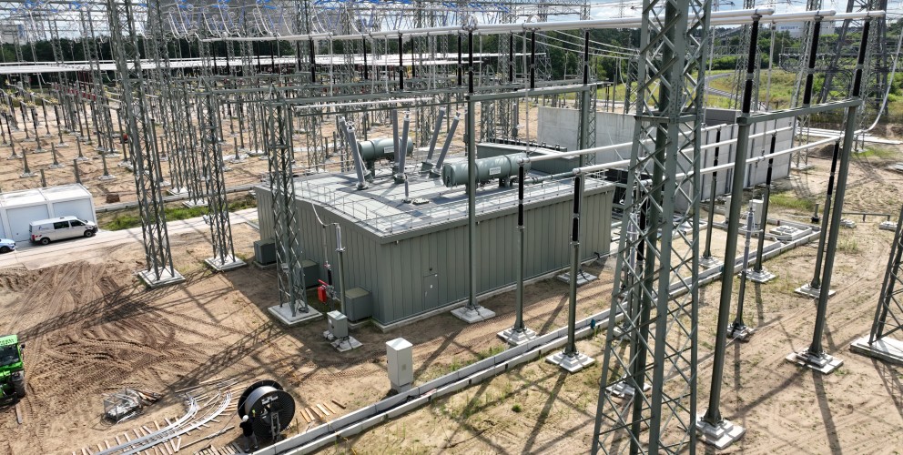 Das Bild zeigt eine großflächige Hochspannungs-Schaltanlage unter freiem Himmel an einem sonnigen Tag mit leicht bewölktem Himmel. Der Boden ist größtenteils sandig-braun und unbewachsen, was auf Bauarbeiten oder eine Freifläche hinweist.