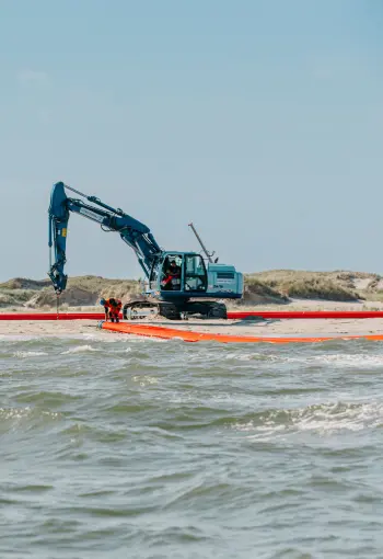 Vordergrund: Die untere Hälfte des Bildes zeigt bewegtes Meerwasser mit kleinen Wellen, die auf den Strand zulaufen.
Mitte: Im flachen Wasser und am Übergang zum Sandstrand verläuft ein langes, leuchtend orangefarbenes Objekt, das wie eine große Ölsperre oder ein flexibles Rohr aussieht. Dieses Objekt erstreckt sich horizontal über die gesamte Breite des Bildes. Zwei Arbeiter in orangefarbener Sicherheitskleidung sind auf der rechten Seite in der Nähe dieses Objekts und des größeren Baggers zu sehen.
Hintergrund: Auf dem Sandstrand stehen zwei große, blaue Hydraulikbagger. Der größere Bagger auf der rechten Seite hat seine Schaufel nach unten gerichtet, möglicherweise für Bauarbeiten oder Wartung. Der kleinere Bagger steht weiter links. Im oberen Bereich des Bildes erstreckt sich eine Dünenlandschaft mit trockener Vegetation unter einem klaren, hellblauen Himmel.