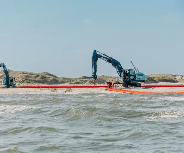 Vordergrund: Die untere Hälfte des Bildes zeigt bewegtes Meerwasser mit kleinen Wellen, die auf den Strand zulaufen.
Mitte: Im flachen Wasser und am Übergang zum Sandstrand verläuft ein langes, leuchtend orangefarbenes Objekt, das wie eine große Ölsperre oder ein flexibles Rohr aussieht. Dieses Objekt erstreckt sich horizontal über die gesamte Breite des Bildes. Zwei Arbeiter in orangefarbener Sicherheitskleidung sind auf der rechten Seite in der Nähe dieses Objekts und des größeren Baggers zu sehen.
Hintergrund: Auf dem Sandstrand stehen zwei große, blaue Hydraulikbagger. Der größere Bagger auf der rechten Seite hat seine Schaufel nach unten gerichtet, möglicherweise für Bauarbeiten oder Wartung. Der kleinere Bagger steht weiter links. Im oberen Bereich des Bildes erstreckt sich eine Dünenlandschaft mit trockener Vegetation unter einem klaren, hellblauen Himmel.