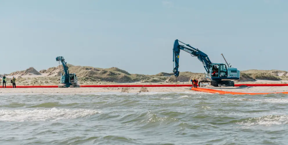 Vordergrund: Die untere Hälfte des Bildes zeigt bewegtes Meerwasser mit kleinen Wellen, die auf den Strand zulaufen.
Mitte: Im flachen Wasser und am Übergang zum Sandstrand verläuft ein langes, leuchtend orangefarbenes Objekt, das wie eine große Ölsperre oder ein flexibles Rohr aussieht. Dieses Objekt erstreckt sich horizontal über die gesamte Breite des Bildes. Zwei Arbeiter in orangefarbener Sicherheitskleidung sind auf der rechten Seite in der Nähe dieses Objekts und des größeren Baggers zu sehen.
Hintergrund: Auf dem Sandstrand stehen zwei große, blaue Hydraulikbagger. Der größere Bagger auf der rechten Seite hat seine Schaufel nach unten gerichtet, möglicherweise für Bauarbeiten oder Wartung. Der kleinere Bagger steht weiter links. Im oberen Bereich des Bildes erstreckt sich eine Dünenlandschaft mit trockener Vegetation unter einem klaren, hellblauen Himmel.