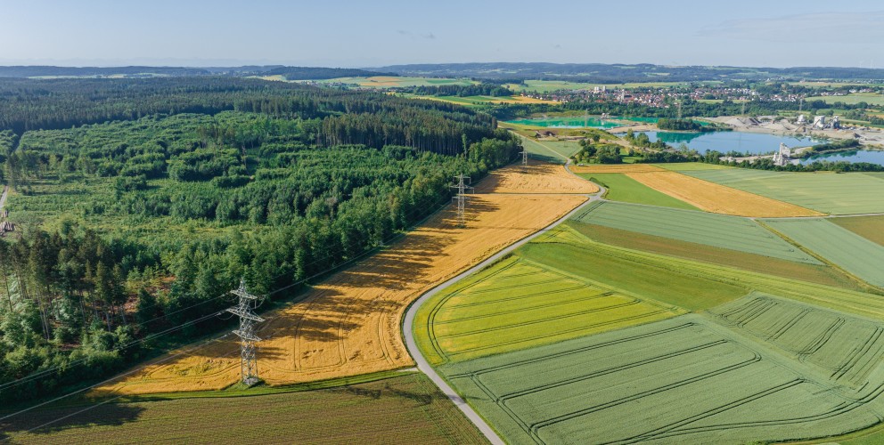 Luftaufnahme einer ländlichen Landschaft mit abwechslungsreichen Feldern in verschiedenen Grüntönen und Gelbtönen, teils bestellt, teils abgeerntet. Ein Strommast mit Hochspannungsleitungen verläuft diagonal durch das Bild und trennt ein Waldgebiet auf der linken Seite von den offenen Feldern rechts. Im Hintergrund sind ein See, ein Kieswerk und eine Ortschaft zu sehen. Die Szene liegt unter klarem, blauem Himmel.