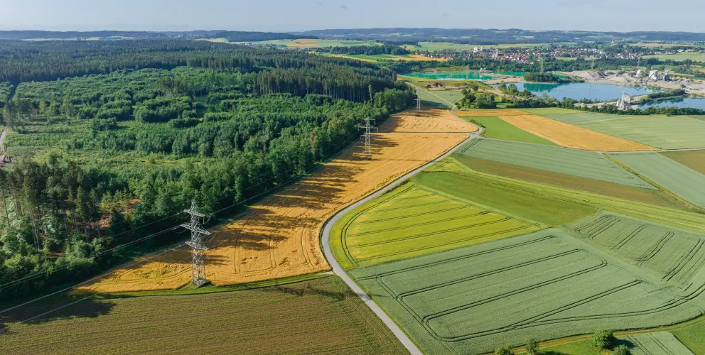 Luftaufnahme einer ländlichen Landschaft mit abwechslungsreichen Feldern in verschiedenen Grüntönen und Gelbtönen, teils bestellt, teils abgeerntet. Ein Strommast mit Hochspannungsleitungen verläuft diagonal durch das Bild und trennt ein Waldgebiet auf der linken Seite von den offenen Feldern rechts. Im Hintergrund sind ein See, ein Kieswerk und eine Ortschaft zu sehen. Die Szene liegt unter klarem, blauem Himmel.