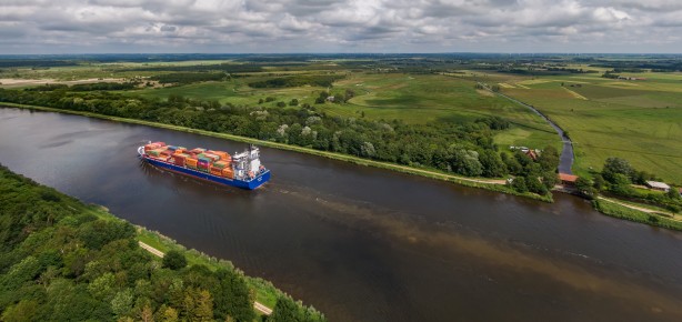 Luftaufnahme eines Containerschiffs auf einem breiten Kanal umgeben von grüner Landschaft und bewölktem Himmel