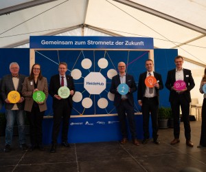 Gruppe von neun Personen steht vor einer blauen Tafel mit dem Schriftzug 'Gemeinsam zum Stromnetz der Zukunft' und hält bunte runde Schilder mit verschiedenen Symbolen wie Stromnetz, Windkraft und Haus