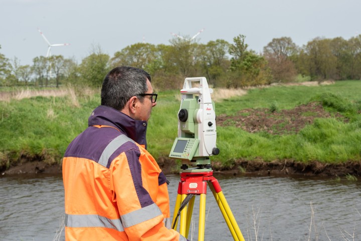 Eine Person in einer orangefarbenen Sicherheitsjacke steht vor einem Messgerät am Ufer eines Gewässers.