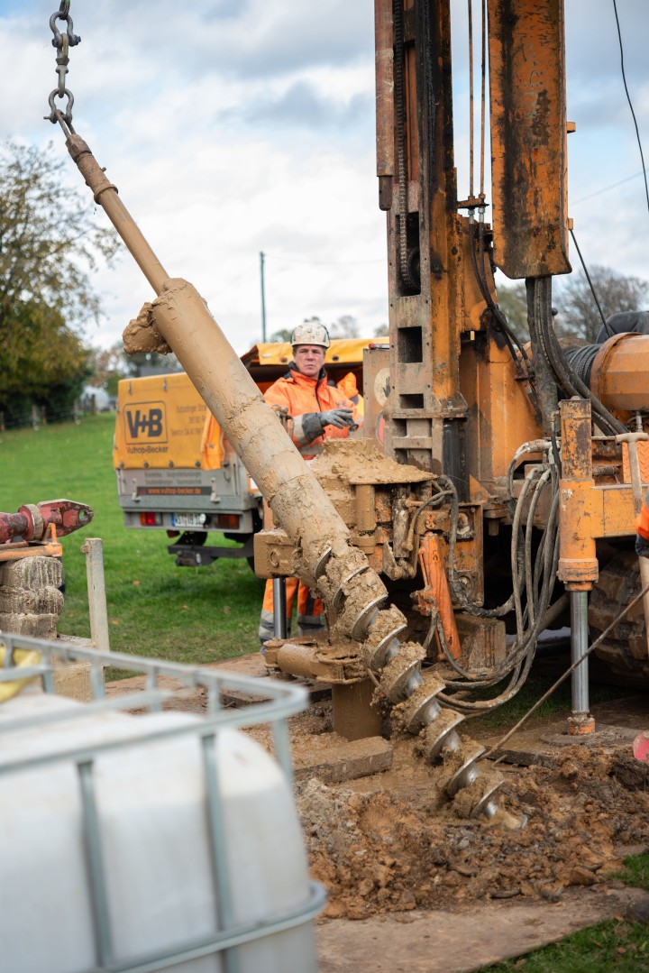 Im Mittelpunkt steht ein großer schneckenförmiger Bohrkopf, der an einem hohen Fahrzeug anfgehägt ist und auf dem Boden stützend schräg zur linken Seite hängt.
