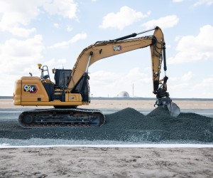 Ein großer gelber Kettenbagger der Firma C&K verteilt mit seiner Schaufel dunklen Schotter oder Kies auf einer Baustelle, im fernen Hintergrund ist die Kuppel eines Kernkraftwerks erkennbar.