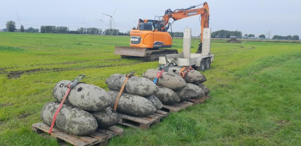 Ein oranger Kettenbagger und ein Anhänger stehen auf einer Wiese hinter vier Holzpaletten, auf denen rund zehn schlammbedeckte Blindgänger mit Spanngurten gesichert sind.