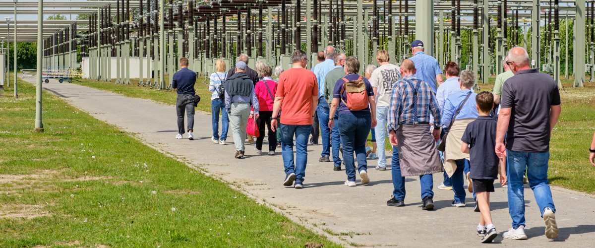 Gruppe von Menschen geht auf einem Weg neben einer großen Anlage mit vielen Metallstreben und Drähten