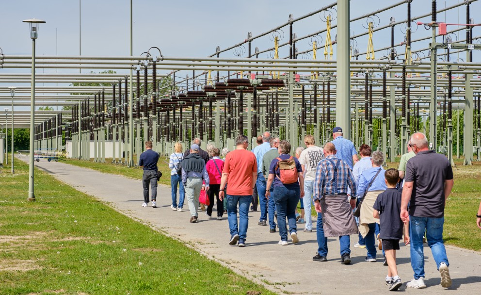 Gruppe von Menschen geht auf einem Weg neben einer großen Anlage mit vielen Metallstreben und Drähten