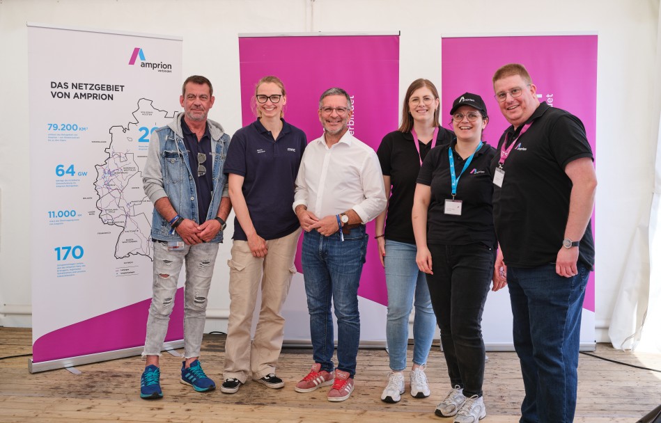 Eine sechsköpfige Gruppe in sportlich-lässiger Kleidung steht nebeneinander und lächelt vor Amprion-Informationsbannern in die Kamera.