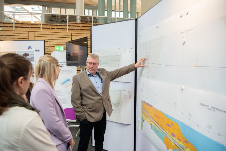 Aufnahme einer Gesprächssituation während einer öffentlichen Informationsveranstaltung: Ein Mann zeigt auf eine Karte und blickt in Richtung zweier Zuhörerinnen.