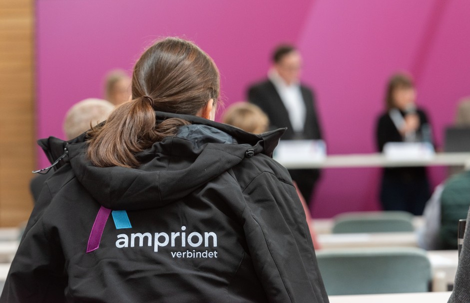 Vortragssitualtion mit Rückenansicht einer Frau in schwarer Jacke mit Amprion-Logo im Vordergrund, im Hintergrund ist eine fuchsiafarbene Wand zu sehen. Davor stehen Menschen an einem Tisch. Der Hintergrund ist verschwommen.