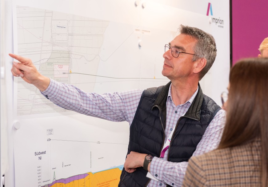 Ein Mann deutet mit dem Zeigefinger auf einen an der Wand hängenden Plan