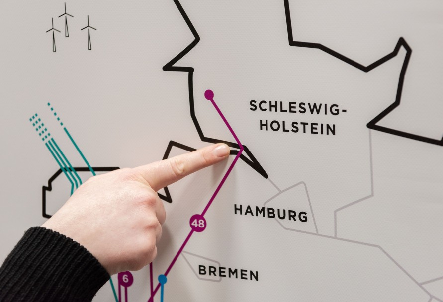 Eine Hand mit ausgestrecktem Zeigefinger deutet auf eine Grafik, auf der die nördlichen Länder Deutschlands und die Luftlinie des Projekts Korridor B dargestellt sind.