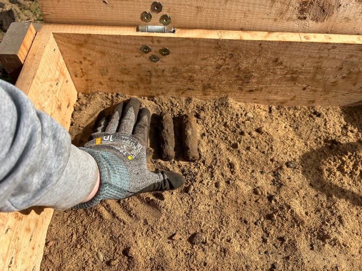 In einer geöffneten Holzkiste mit braunem Sand liegen zwei Stangen, die rund und fingerlang sind. Eine Hand mit Handschuh liegt daneben.