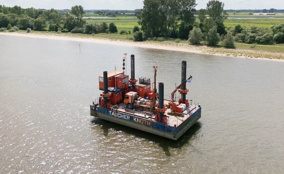Eine orange-rote Arbeitsplattform der Firma Taucher Knoth ist auf einem breiten Fluss vor einem grünen Ufer verankert.