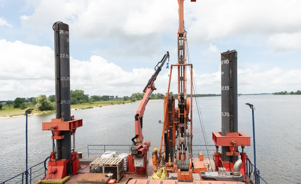 Eine orange-schwarze Arbeitsplattform mit Bohr- und Kranvorrichtungen sowie Markierungspfählen führt Arbeiten auf einem Fluss oder Gewässer durch.