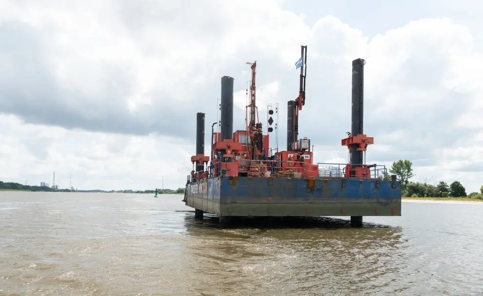 Eine rote und blaue Arbeitsplattform mit vier hohen Pfählen ankert auf einem breiten Fluss unter einem bewölkten Himmel.