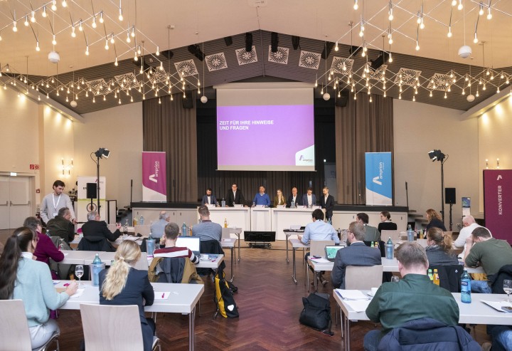 Informationsveranstaltung für Träger öffentlicher Belange am 27. November 2024: Zu sehen sind sieben Personen, die hinter drei großen weißen Tischen vor einer Leinwand stehen. Mehrere Tischreihen sind im Vordergrund aufgebaut, daran sitzen etwaige Personen mit dem Rücken zur Kamera.