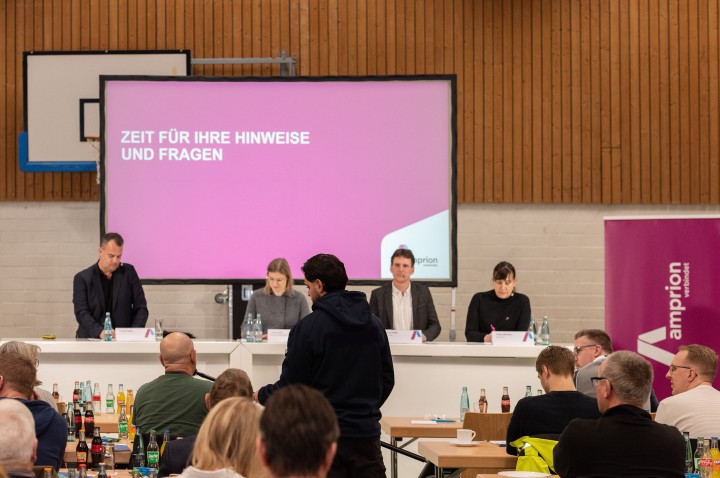 Informationsveranstaltung für Träger öffentlicher Belange am 25. Januar 2025: Zu sehen sind vier Personen, die hinter drei großen weißen Tischen vor einer Leinwand stehen. Mehrere Tischreihen sind im Vordergrund aufgebaut, daran sitzen etwaige Personen mit dem Rücken zur Kamera. Zwischen den Tischreihen steht eine Person, die einer anderen sitzenden Person ein Mikrofon hinhält.