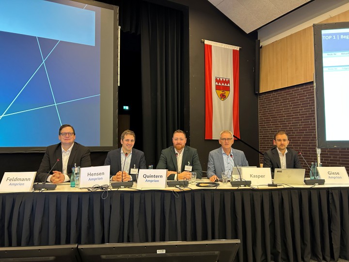 Fünf Männer im Anzug sitzen an einem langen Tisch, der vorne mit Stoff verkleidet ist. Auf dem Tisch stehen Namensschilder. Alle blicken in Richtung Kameralinse.