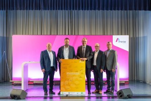 Das Bild zeigt fünf Männer in Anzügen, die gemeinsam vor einer pinkfarbenen Kulisse mit dem Amprion-Logo und einem orangefarbenen Schild posieren. Die Szene ist ein offizieller Fototermin im Rahmen einer Pressekonferenz oder Veranstaltung.
