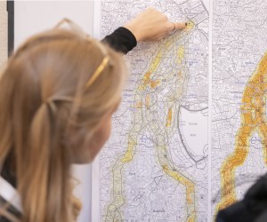 Vordergrund: Im linken Teil des Bildes ist der Hinterkopf und die rechte Profilseite einer Frau mit schulterlangem, blondem Haar zu sehen, das in zwei Zöpfen zusammengebunden ist. Sie trägt eine goldfarbene Brille.
Hintergrund und Aktion: Der Fokus liegt auf der rechten Hand der Frau, die mit dem Zeigefinger auf einen spezifischen, gelb markierten Bereich auf einer großformatigen, technischen Karte deutet. Die Karte zeigt komplexe Linienführungen, Höhenlinien und geografische Markierungen.