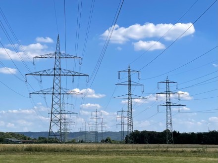 Fotografie einer ländlichen Ebene mit mehreren parallel verlaufenden Reihen von Hochspannungsmasten unterschiedlicher Bauart unter einem leicht bewölkten, blauen Himmel.