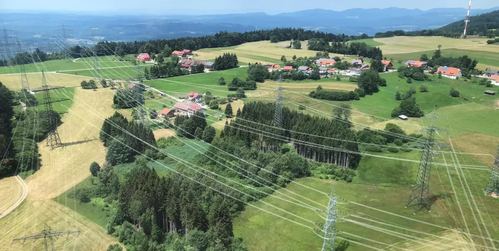 Eine Luftaufnahme im Querformat zeigt eine weitläufige, hügelige Kulturlandschaft, durchzogen von mehreren Hochspannungsfreileitungen und ihren Masten.
Vordergrund: Dichter Mischwald bedeckt den unteren Bereich, gefolgt von landwirtschaftlichen Flächen (grüne Wiesen und gelbe Felder). Mehrere filigrane Gittermasten ragen aus den Feldern heraus.
Mitte/Hintergrund: In der Ferne sind verstreute Gehöfte und kleine Siedlungen mit rot gedeckten Häusern inmitten von Wäldern und Feldern sichtbar. Eine größere Waldfläche befindet sich im Zentrum der Aufnahme.
Skyline: Am Horizont zeichnen sich dunklere Umrisse von Bergen oder Hügelketten unter einem bewölkten Himmel ab.
Perspektive: Die Aufnahme wurde vermutlich aus großer Höhe (z. B. aus einem Flugzeug, Hubschrauber oder von einem hohen Turm) aufgenommen, was die vielen parallelen Stromleitungen über das gesamte Bild hinweg sichtbar macht.