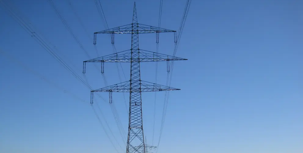 Das Bild zeigt einen einzelnen mast für Hochspannungsleitunge. Er steht in einer grünen, offenen Landschaft, wahrscheinlich ein Feld oder eine Wiese.
Leitungen:
Vom Mast aus erstrecken sich mehrere Hochspannungsleitungen in verschiedene Richtungen. Sie verlaufen in einem leichten Durchhang zwischen den Masten, die außerhalb des Bildausschnitts liegen.
Umgebung:
Der Mast steht auf einer grünen Fläche mit kurzem Gras. Im Hintergrund ist eine Baumreihe zu sehen, die den Horizont bildet. Der Himmel ist klar und blau, ohne Wolken. Das Licht deutet auf einen sonnigen Tag hin.